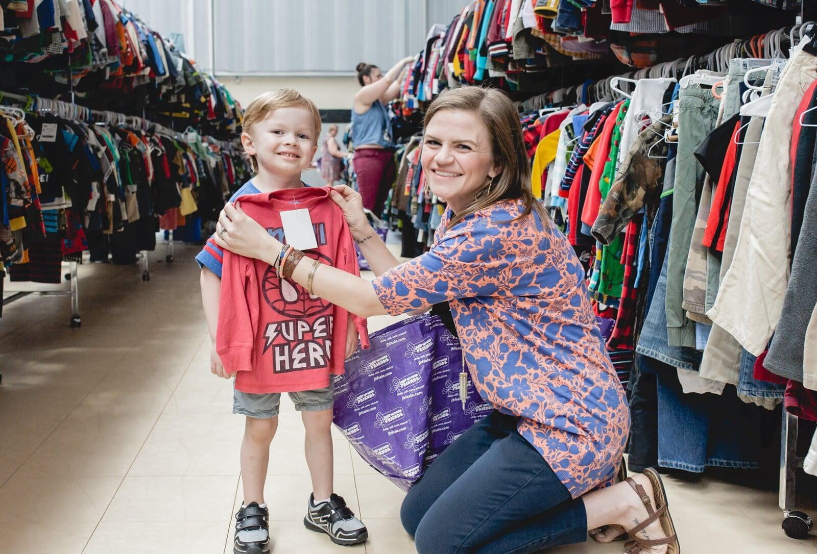 A mother and son stand in a clothing aisle while mom holds up a shirt to see if it will fit son.