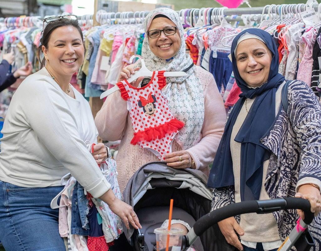 Three women stand around a baby in a stroller, holding clothing they are shopping for at the sale.